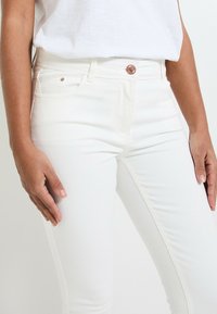 Vaqueros de mezclilla blanca con un ajuste slim, con cierre de botón de cobre y bolsillos frontales. Textura suave y detalles de costura limpios.
