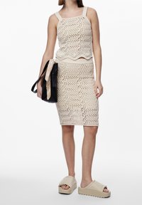 Top de crochet de punto beige con tirantes anchos y dobladillo festoneado, combinado con una falda a la altura de la rodilla a juego, complementado con sandalias de plataforma beige y un bolso negro.