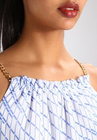 Il volto inferiore e il collo di una donna che indossa un top bianco con un motivo a rombi blu e spalline in oro, labbra truccate con rossetto di un rosso scuro.