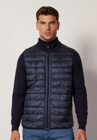 Granatowa kamizelka puchowa z poziomymi przeszyciami, wysokim kołnierzem i pełnym zamkiem błyskawicznym. Noszona na ciemnej bluzce, zaprezentowana na neutralnym tle.