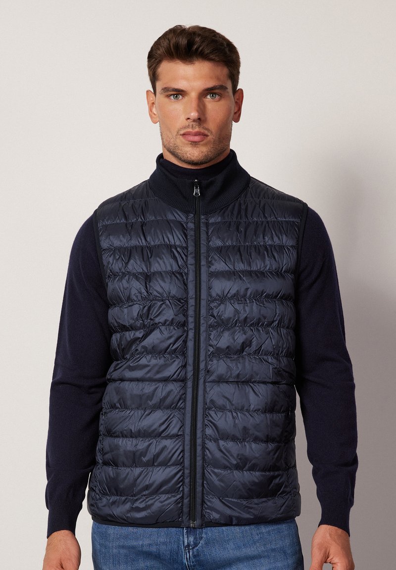 Granatowa kamizelka puchowa z poziomymi przeszyciami, wysokim kołnierzem i pełnym zamkiem błyskawicznym. Noszona na ciemnej bluzce, zaprezentowana na neutralnym tle.