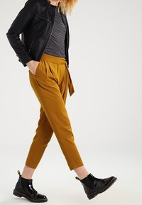 Schwarze Lederjacke, gestreiftes T-Shirt, senfgelbe, taillierte Hose mit gebundenem Bund und glänzende schwarze Chelsea-Boots mit einem gelben Etikett.