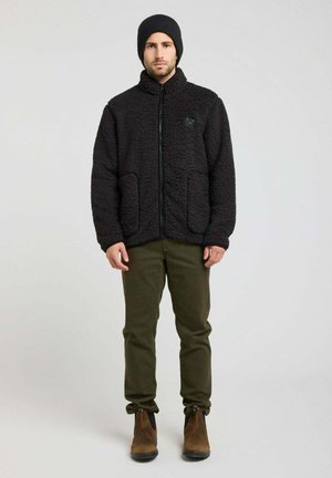 Homme portant une veste polaire noire, un bonnet noir, un pantalon vert olive et des bottines marron, debout devant un fond clair uni.