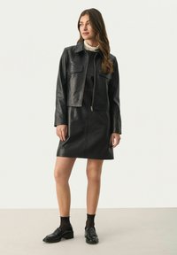 Schwarze Lederjacke mit einem cropped Design, ausgestattet mit zwei Fronttaschen. Kombiniert mit einem passenden Lederrock. Das Model trägt schwarze Schlappen und Socken.