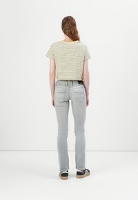Kuttet gul- og hvitstripet t-skjorte, grå slim-fit jeans og svarte sneakers med hvite striper. Enfarget bakgrunn, sett fra baksiden.