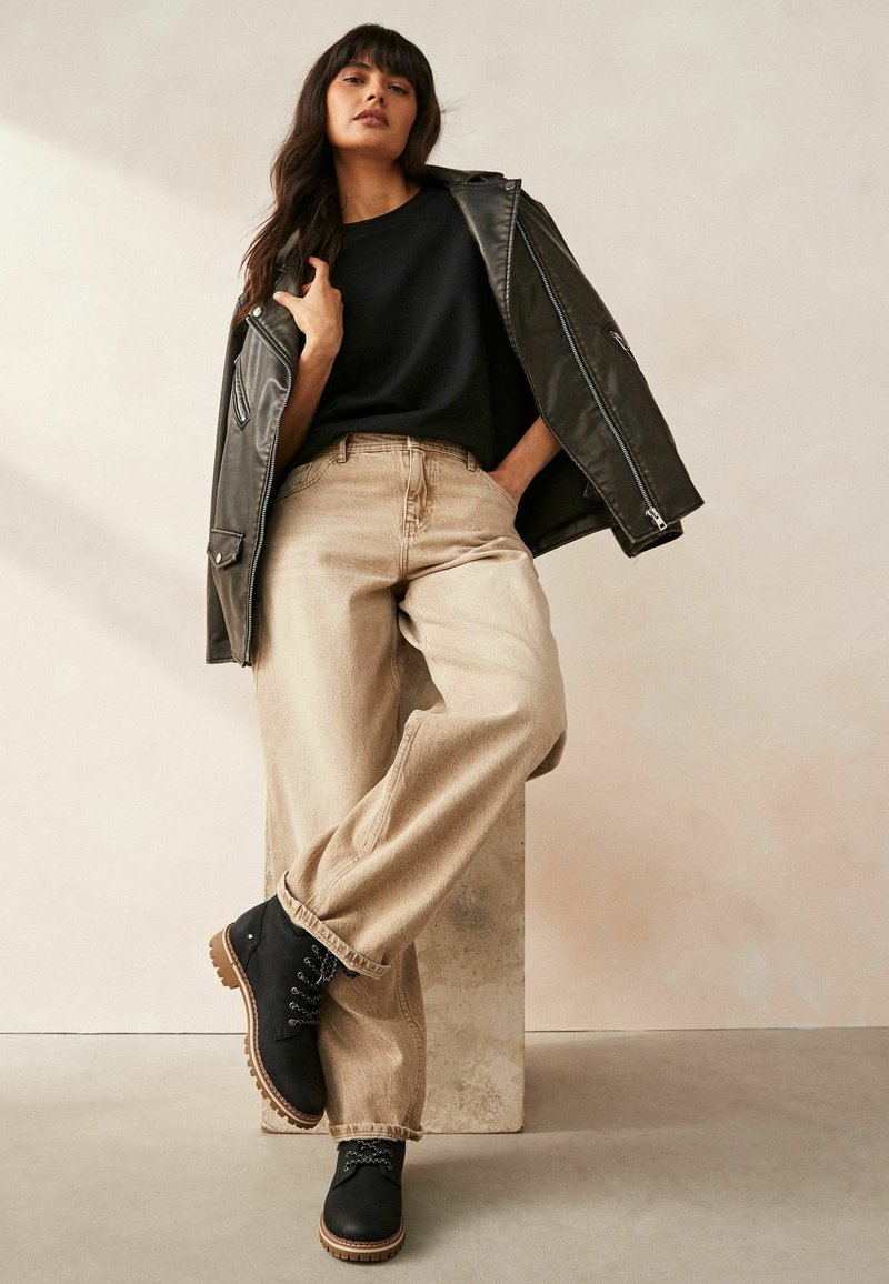 Chaqueta de cuero negra, vaqueros beige holgados y botas negras con cordones y suelas texturizadas. Top negro simple. De pie contra un fondo neutro.
