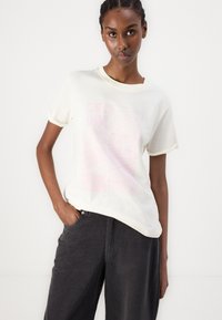 Cremfarbendes T-Shirt mit kurzen Ärmeln und lockerer Passform, mit einem pinken Grafikdesign. Kombiniert mit hochtaillierten dunkelgrauen Hosen.