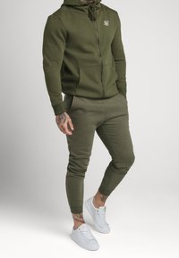 Olijfgroene hoodie met ritssluiting en trekkoorden, bijpassende joggingbroek en witte sneakers. Voorzien van een logo op de borst en aansluitende manchetten.