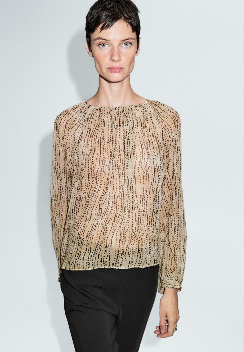 Durchsichtige Bluse mit langen Ärmeln und runder Ausschnitt in Beige, mit einem strukturierten, wellenförmigen Muster in dunkleren Tönen. Kombiniert mit schwarzen Hosen.