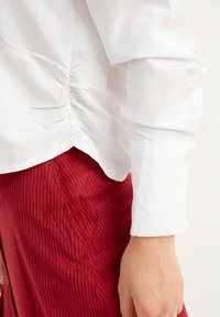 Blusa bianca con dettagli arricciati sui lati, abbinata a una gonna di velluto a coste rossa con righe verticali e orlo leggermente svasato.