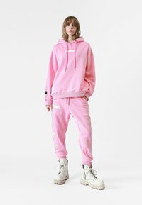 Sudadera de algodón rosa con cordones y un parche en el pecho, combinada con joggers a juego que presentan acentos texturizados y parches bordados en blanco.
