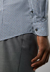 Langarmhemd in Hellblau mit geometrischem Muster. Mit kontrastierenden Knöpfen, eng anliegenden Manschetten und glattem Stoff. Graue Hose sichtbar.