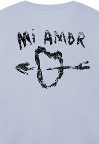 Lichtblauw T-shirt met een zwarte afbeelding van een hart doorboord door een pijl, met de tekst "Mi Amor" bovenaan in een vet, schilderachtig lettertype.
