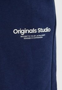 Tessuto della felpa blu navy con texture a coste. Presenta un testo stampato bianco: "Originals Studio" e un ulteriore marchio sottostante.
