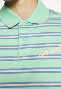 Gros plan d'un polo vert à col avec des rayures horizontales bleues et rose clair et deux boutons verts sur la patte de boutonnage.