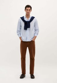 Camisa de botones rayada en azul claro, suéter azul marino atado al cuello, pantalones slim-fit marrones y zapatos negros. Fondo plain.