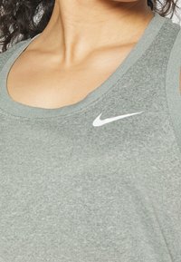 Šedý sportovní tílko s širšími ramínky, hladkým povrchem a bílým logem Nike swoosh na hrudi. Jednoduchý design, volnočasový styl.