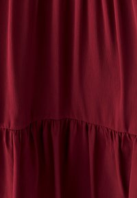 Bordo suknelė su tekstūriniu, sluoksniuotu dizainu. Yra glotni viršutinė dalis ir susiraukšlėjusios raukiniai apatinėje dalyje. Minkšta, lengva medžiaga.