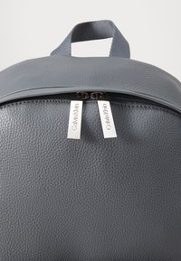Sac à dos en cuir texturé gris avec une poignée supérieure et deux curseurs de fermeture éclair argentés gravés des logos Calvin Klein.