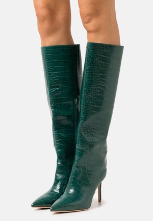 Bottes vert foncé en cuir à motif crocodile, montant jusqu'au genou, avec bouts pointus et talons aiguilles, portées sur des jambes nues.
