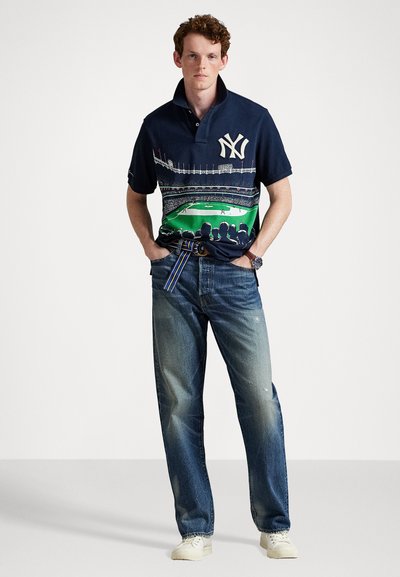 Ναυτικό πόλο μπλουζάκι με κεντημένο λογότυπο, που διαθέτει γραφικό σχέδιο ενός γηπέδου μπέιζμπολ. Φοριέται με ξεθωριασμένο μπλε τζιν και λευκά αθλητικά παπούτσια.