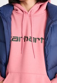 Person som bär en rosa Carhartt-hoodie med snörningar och en marinblå vadderad jacka, beskuren för att endast visa torso och nacke.