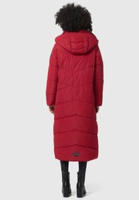 Red puffer coat with a hood, featuring chevron stitching, a long length, and side pockets. Worn over black leggings and heeled boots.
