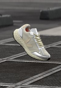 Białe i srebrne sneakersy z teksturowanym siateczkowym wierzchem, okrągłym czubkiem i grubą, wzorzystą podeszwą; mają beżowy akcent po bokach.