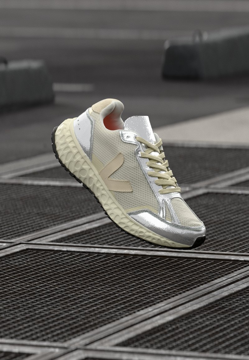 Białe i srebrne sneakersy z teksturowanym siateczkowym wierzchem, okrągłym czubkiem i grubą, wzorzystą podeszwą; mają beżowy akcent po bokach.
