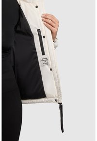 Biała kurtka puchowa z gładkim wykończeniem, czarną podszewką, kieszenią na zamek i metką wewnątrz z napisem „Ciepłe objęcie zrobione z tkaniny.”