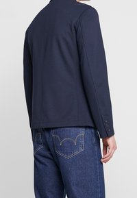 Námořnický modrý blejzr, přiléhavý střih, hladká látka, dlouhé rukávy s manžetami na čtyři knoflíky, nošený přes džíny s vzorovaným prošíváním na kapsách.