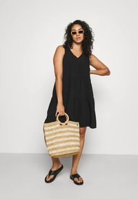 Musta hihaton mekko, jossa on V-kaula-aukko ja kerroksittainen muotoilu, yhdistettynä kudottuun beigeen ja kermaan raidalliseen laukkuun. Mallilla on mustat varvassandaalit.