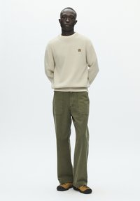 Sweat-shirt beige clair avec encolure ronde et petit logo. Pantalon large kaki avec poches avant. Chaussures noires et orange.