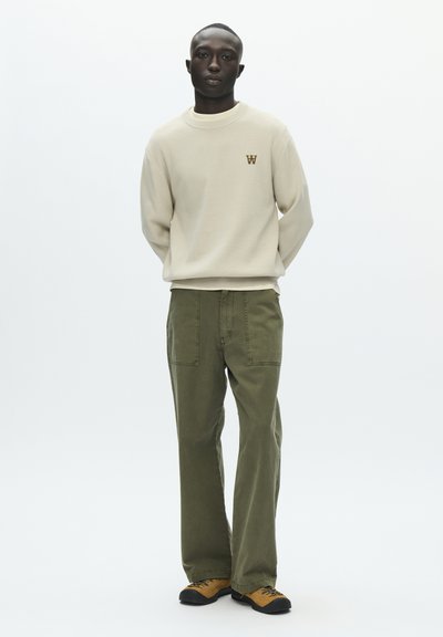 Sweat-shirt beige clair avec encolure ronde et petit logo. Pantalon large kaki avec poches avant. Chaussures noires et orange.
