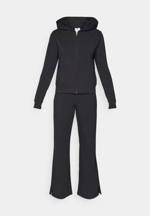 Felpa nera con zip, cappuccio e tasche frontali abbinata a pantaloni neri aderenti con spacchi laterali alle caviglie, realizzati in tessuto morbido.
