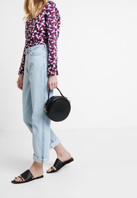 Sac à main circulaire noir avec une poignée, jean bleu clair roulé, sandales noires à enfiler, et un haut à manches longues à motif géométrique coloré.