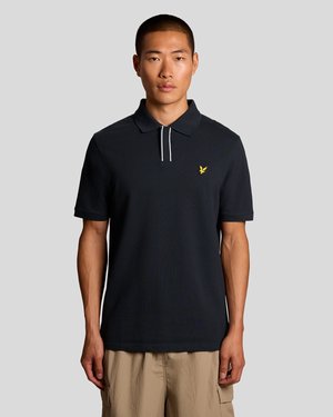 Jeune homme portant un polo à manches courtes noir avec un petit logo d'oiseau jaune sur la poitrine et un pantalon cargo beige.