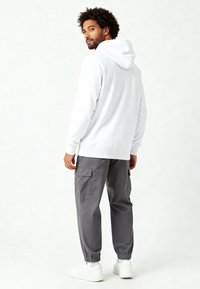 Felpa bianca con cappuccio e polsini elasticizzati, abbinata a pantaloni cargo grigi con tasche laterali e scarpe da ginnastica bianche. Texture liscia.