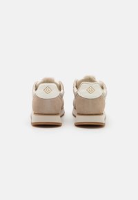 Ein Paar beigefarbene Wildledersneaker, die von hinten gezeigt werden. Sie verfügen über weiße Lederfersenpatches mit einem goldenen "G"-Logo auf weißem Hintergrund.