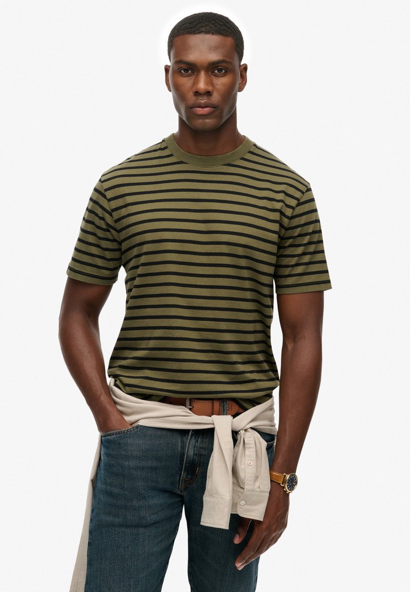 Olive green striped short-sleeve t-shirt with black stripes, round neckline, paired with dark blue jeans and a cream sweater tied at the waist.