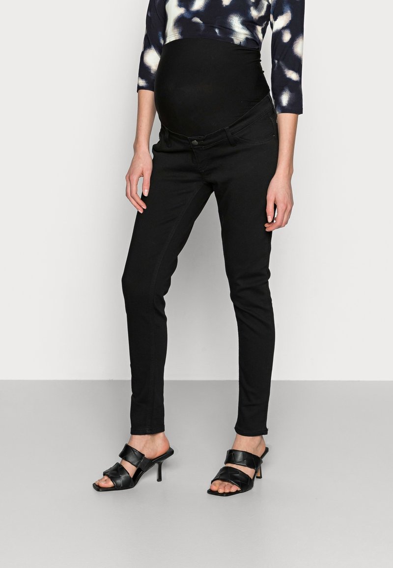 Persona embarazada vestida con pantalones de maternidad negros con panel elástico en la barriga, una blusa de tie-dye y sandalias negras de tacón, de pie sobre un suelo gris.