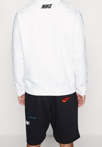 Vit sweatshirt med ribbade ärmslut, med en svart Nike-logotyp på baksidan. Kombinerad med svarta shorts med röda och blå accenter.