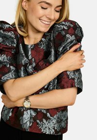 Gornji dio s cvjetnim uzorkom i naboranim rukavima u tamnocrvenoj i srebrnoj boji, s teksturiranim materijalom. Model prikazuje dizajn držeći se za tijelo.