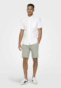 Witte korte mouwen button-up shirt met een borstzak, gecombineerd met lichtgrijze shorts. Draagbaar met witte sneakers met een zwart logo.