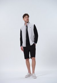 Lys grå ærmeløs jakke med sorte detaljer, parret med en sort skjorte og sorte shorts. Modellen bærer lyse sneakers.