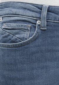 Gros plan sur un jean en denim bleu montrant la poche avant, la petite poche à monnaie, la boucle de ceinture, les coutures et le détail des rivets métalliques.