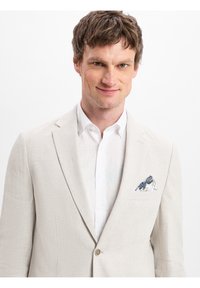 Licht beige linnen blazer met een notch lapel, enkele knoopsluiting en een wit shirt eronder. Voorzien van een blauw-wit patroon zakdoekje.