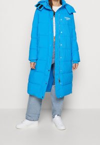 Blå puffjacka med huva, med dragkedja och tryckknappsstängning, sydda paneler och en logo på bröstet. Bärs över avslappnade jeans.