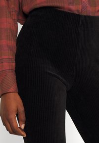 Svarta corduroybyxor med vertikal ribbing, hög midja och slät textur, i kombination med en rutiga skjorta med röda och gröna toner.