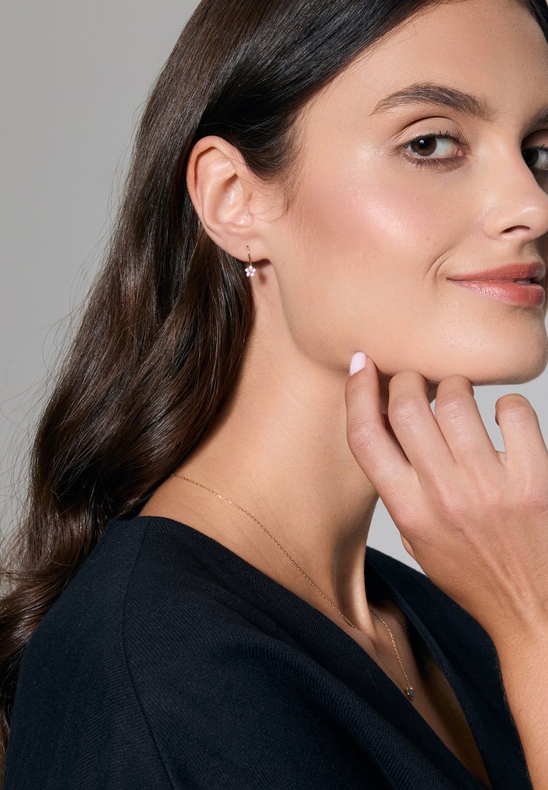 Złote kolczyki w kształcie kół z małym kwiatowym akcentem. Modelka ma długie, ciemne włosy, nosi czarną bluzkę na neutralnym tle.